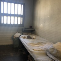 Concrete room with a single bed, dirty bedding, and a few possessions in front of a barred window