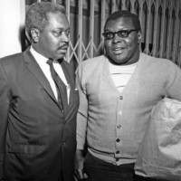 Older man in suit and and younger man in sweater stand in front of a gate