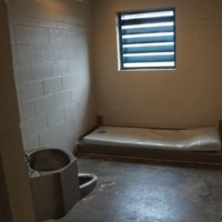 Small empty cell with single bed frame, mattress, sink, toilet, bright overhead light, and barred window