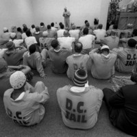 About forty detainees sit on the floor observing one man standing and holding a book