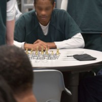 A prisoner sits in front of a chess board, others observe the game