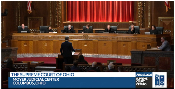 The seven justices of the Supreme Court of Ohio seated at the bench during an oral argument, with an attorney standing at the lectern addressing the court, in the ornate wood-paneled courtroom of the Moyer Judicial Center in Columbus, Ohio.